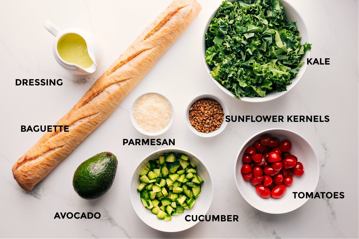 Ingredients In Kale And Avocado Salad All the ingredients in this recipe prepped out for easy assembly including the dressing, baguette, avocado, cucumber, parmesan, sunflower kernels, and tomatoes.