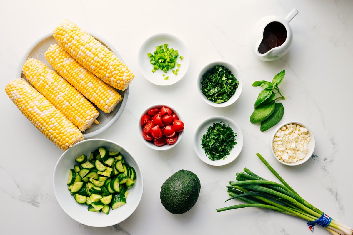 Ingredients In This Corn Salad Recipe All the ingredients in this recipe prepped out for easy assembly including the corn, cucumbers, avocado, tomatoes, jalapeños, cilantro, parsley, basil, green onions, feta, and vinaigrette.