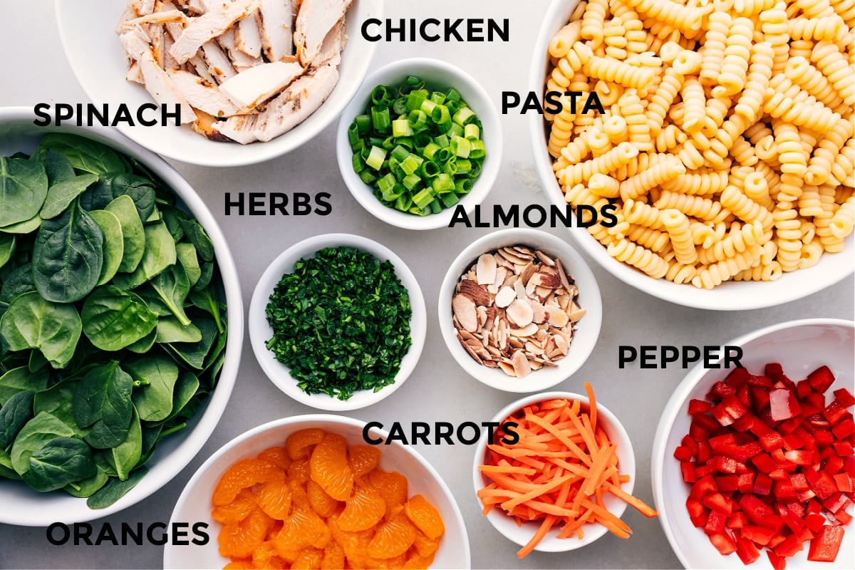 Asian pasta salad ingredients laid out for easy assembly including chicken, pasta, almonds, herbs, spinach, peppers, carrots, and oranges.