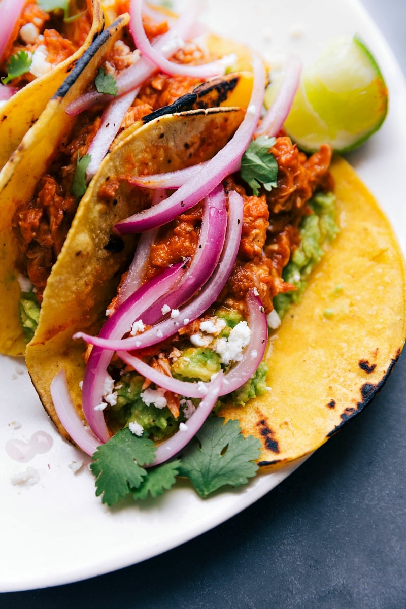 Chicken tinga tacos fully assembled and ready to be enjoyed, topped with red onions, cilantro, cheese, and more.