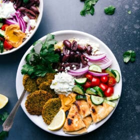 Baked falafel served on a plate with pita bread, tzatziki sauce, olives, onions, tomatoes, and cucumbers.