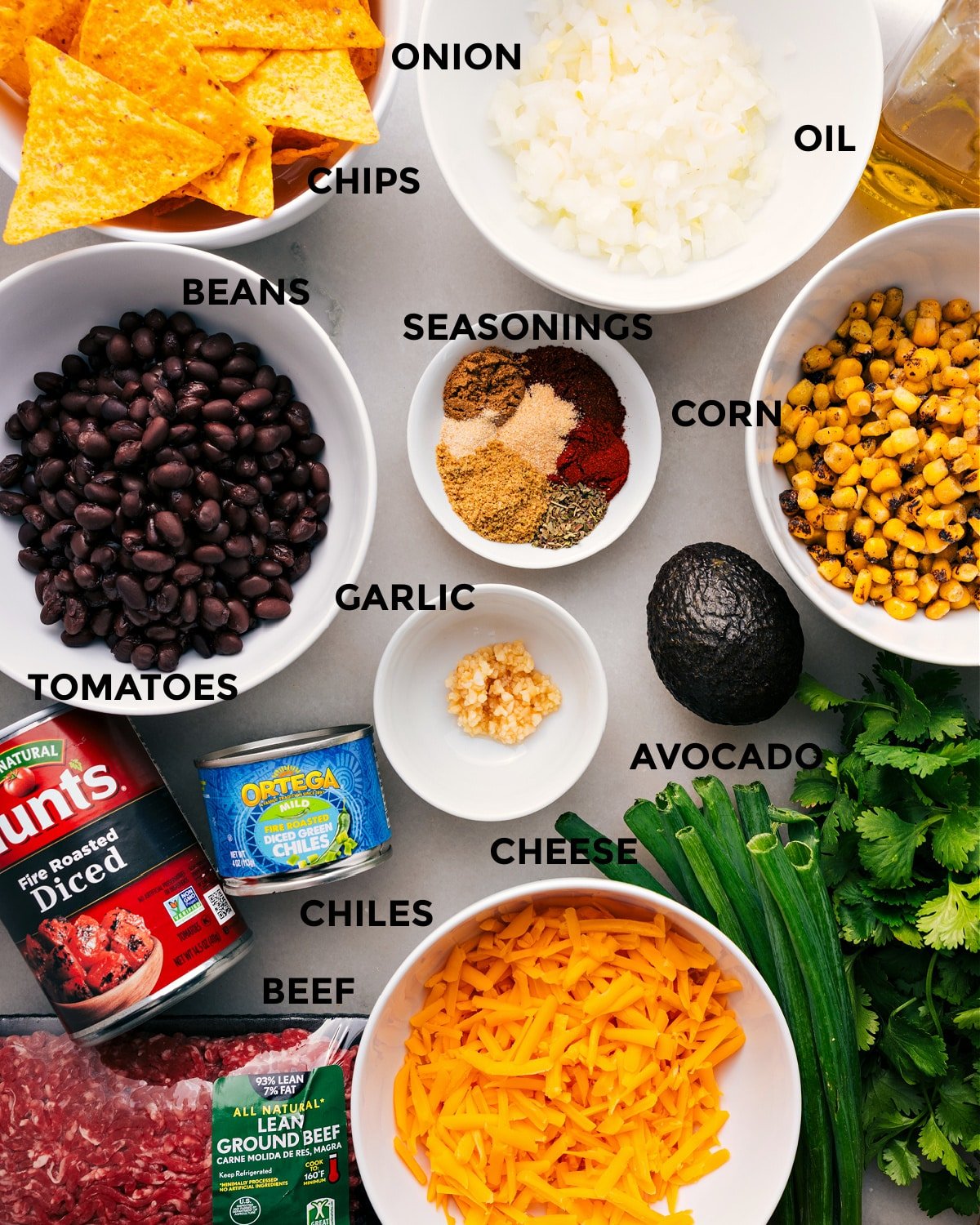 Prep all the ingredients for easy assembly, including the seasonings, meat, corn, avocado, garlic, tomatoes, chiles, cheese, chips, and fresh herbs.