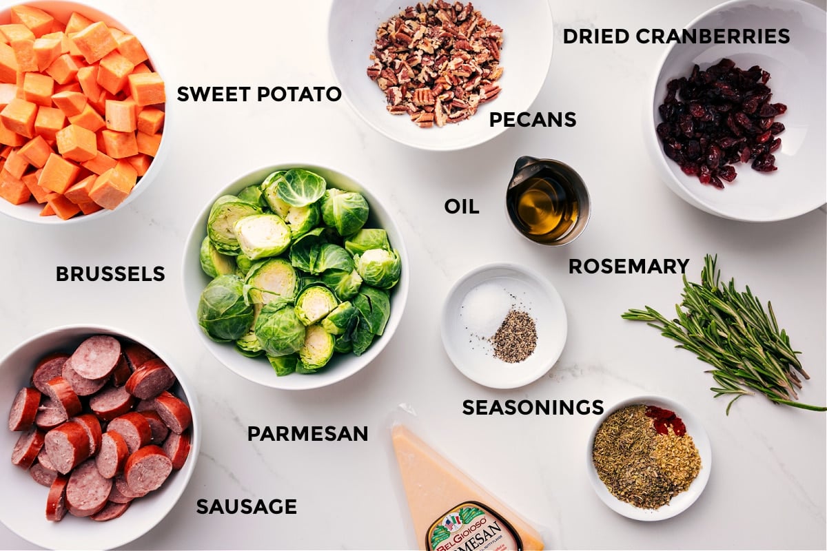 Prepped ingredients for sheet pan: sweet potatoes, sausage, Brussels sprouts, rosemary, cranberries, pecans, Parmesan.