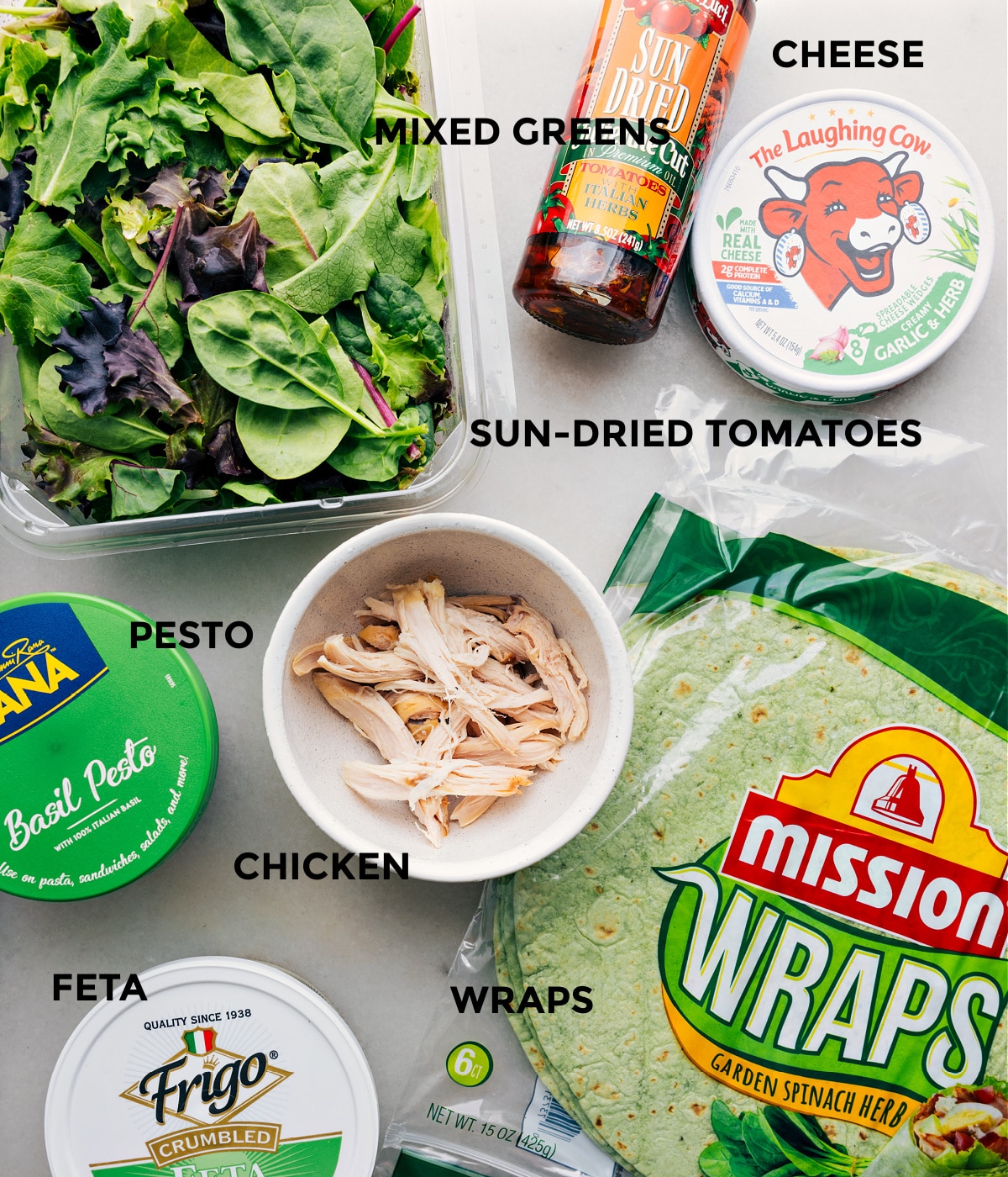 All the ingredients in this recipe prepped out for easy assembly, including pesto, chicken, mixed greens, cheese, tortillas, and feta.
