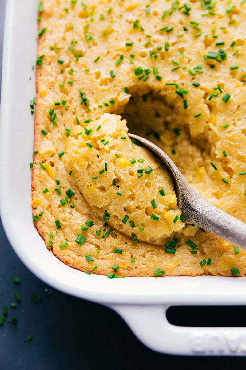 This hearty and flavorful Corn Casserole is made from scratch and is a wonderful addition to your holiday table. It's thick, moist, and the perfect balance of sweet and savory. Recipe via chelseasmessyapron #recipe #cream #baked #sweet #easy #withoutjiffy #thanksgiving #best #quick #simple #chives #sidedish #corn #casserole Overhead image of Corn Casserole with a spoonful being taken out of it