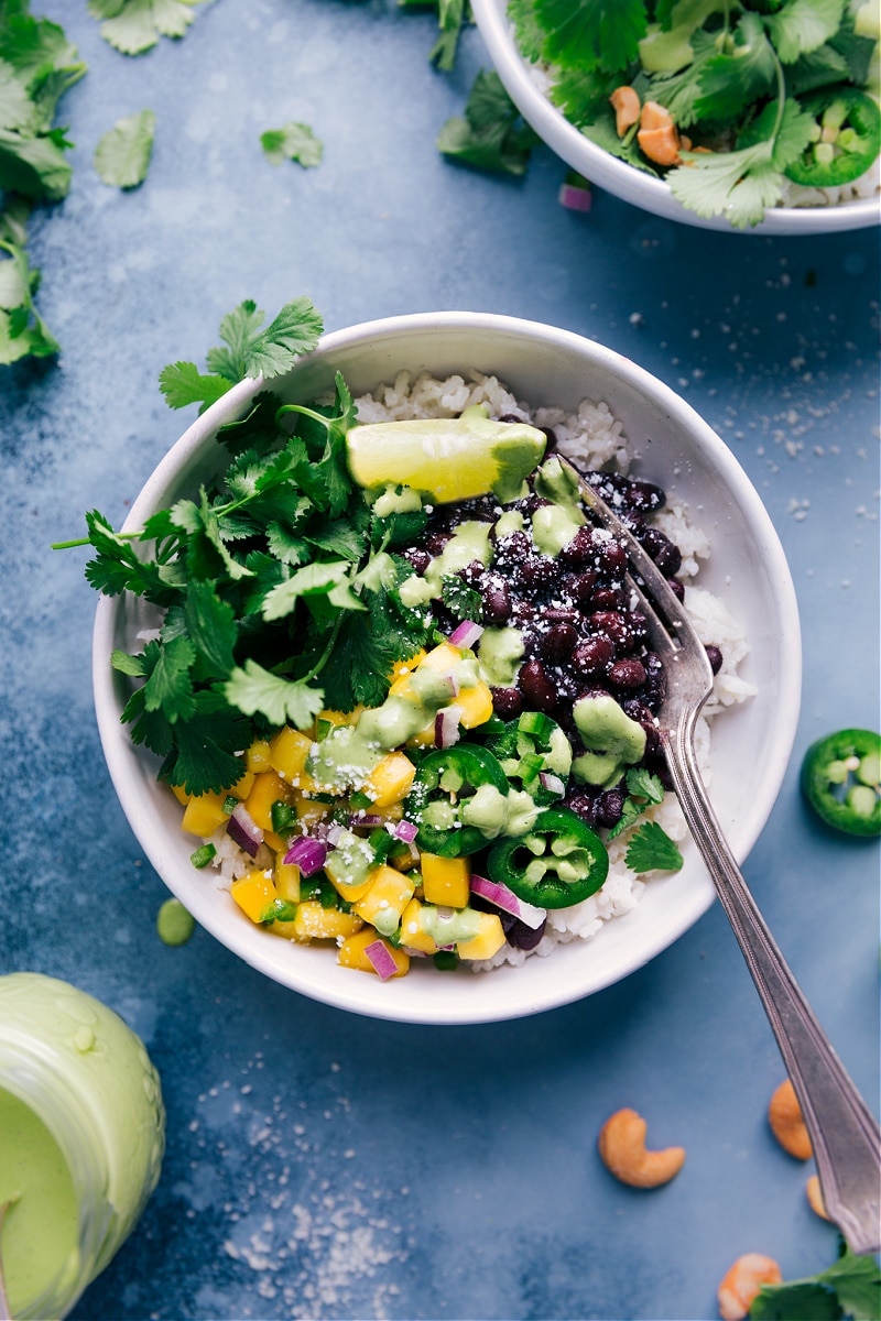 These saucy Black Bean Bowls are one of our new favorite vegetarian meals! They start with coconut-lime rice and are loaded up with ultra-flavorful and saucy black beans and a quick mango-jalapeño topping. A simple (and optional) cashew-cilantro sauce gets drizzled on everything, adding a citrusy freshness. Recipe via ChelseasMessyApron.com #blackbeanbowl #beans vegetarian #citrus #coconutrice Overhead view of Black Bean Bowls with all the toppings.
