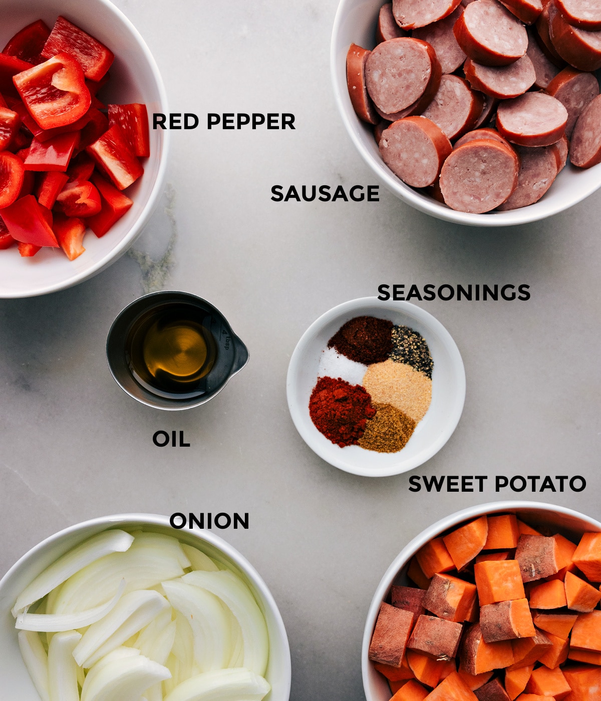 Assorted raw ingredients for cooking arranged on a tray, including sausage, chopped vegetables, olive oil, and seasonings.