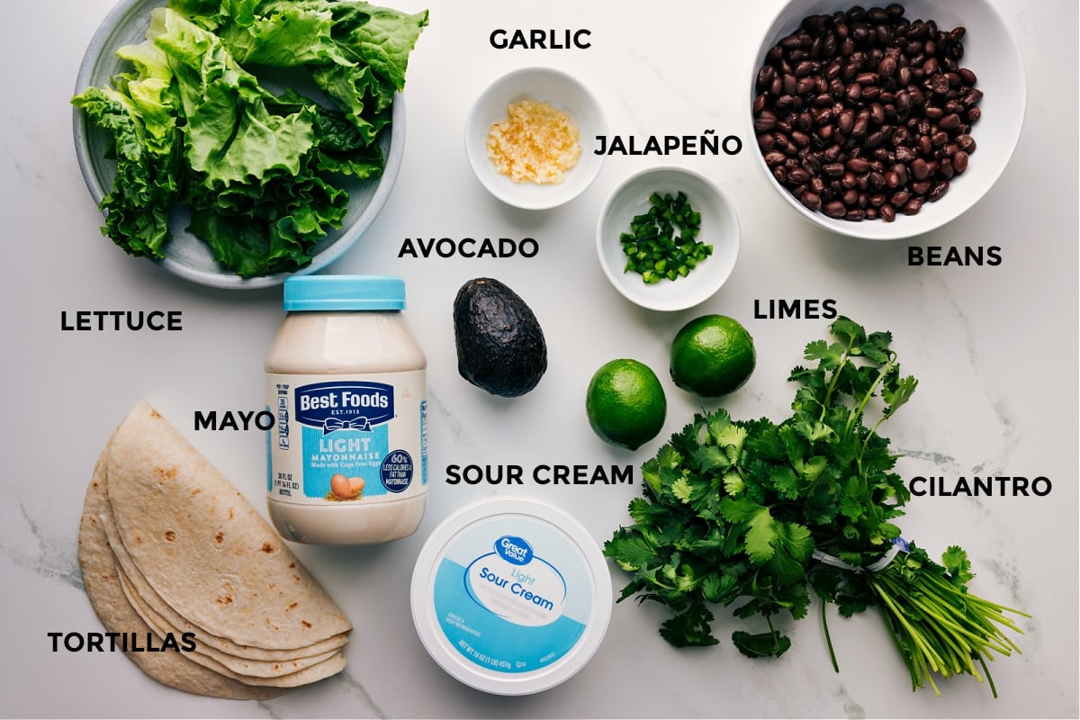 All the ingredients for this recipe prepped and ready for easy assembly, including lettuce, avocado, limes, beans, sour cream, mayo, cilantro, and tortillas.