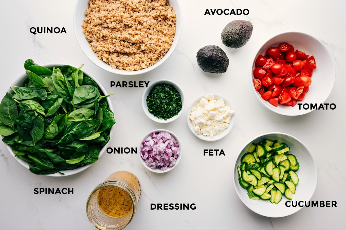 Ingredients In Quinoa Salad All the ingredients prepped for easy assembly, including quinoa, avocado, tomato, feta, cucumber, parsley, onion, dressing, and spinach.