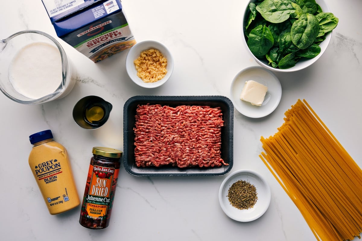 All the ingredients in this recipe prepped out for easy assembly including the cream, dijon, sun-dried tomatoes, broth, garlic, sausage, seasonings, spinach, butter, and pasta.