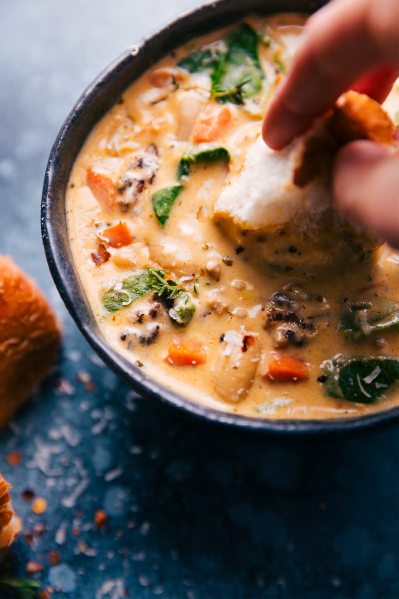 Hearty White Bean and Sausage Soup! This is the best soup! Recipe via ChelseasMessyApron #dinner #quick #easy #simple #spinach #carrots #bread #comfortfood #kidfriendly #familyfriendly #whitebean #soup Image of a piece of bread being dipped into the soup