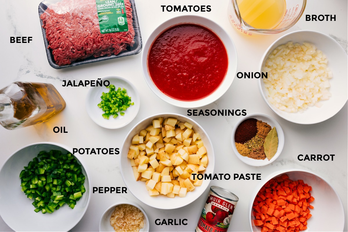 All the ingredients for this recipe prepped and ready for easy assembly, including the beef, broth, tomatoes, onions, seasonings, carrot, tomato paste, potatoes, jalapeño, oil, pepper, and garlic.