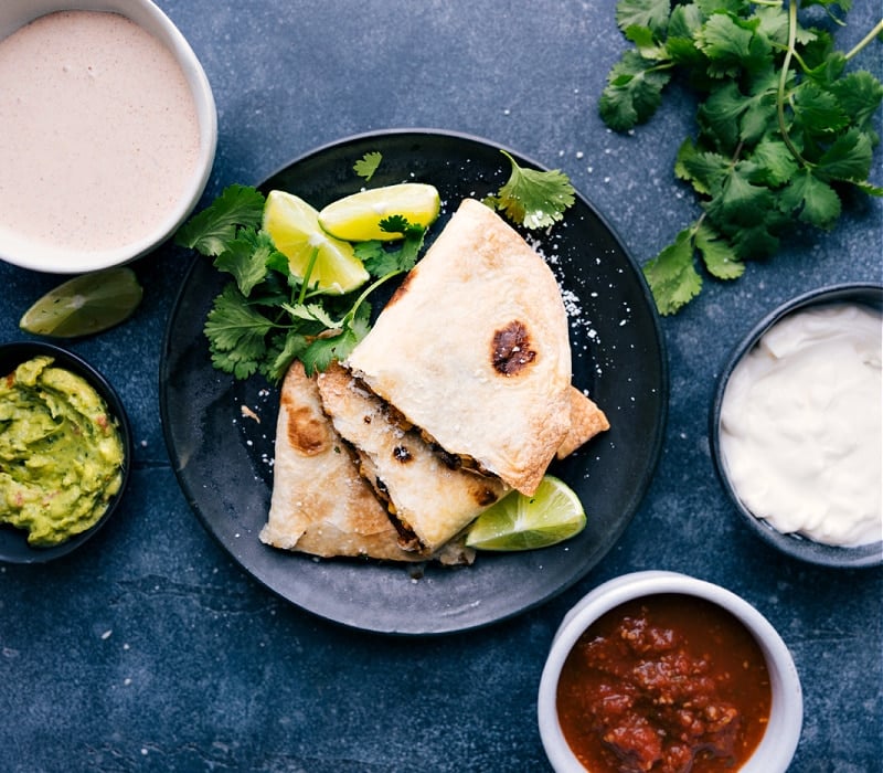 View of Black Bean Quesadilla triangles, with sour cream, salsa, guacamole, chili lime sauce and cilantro on the side.