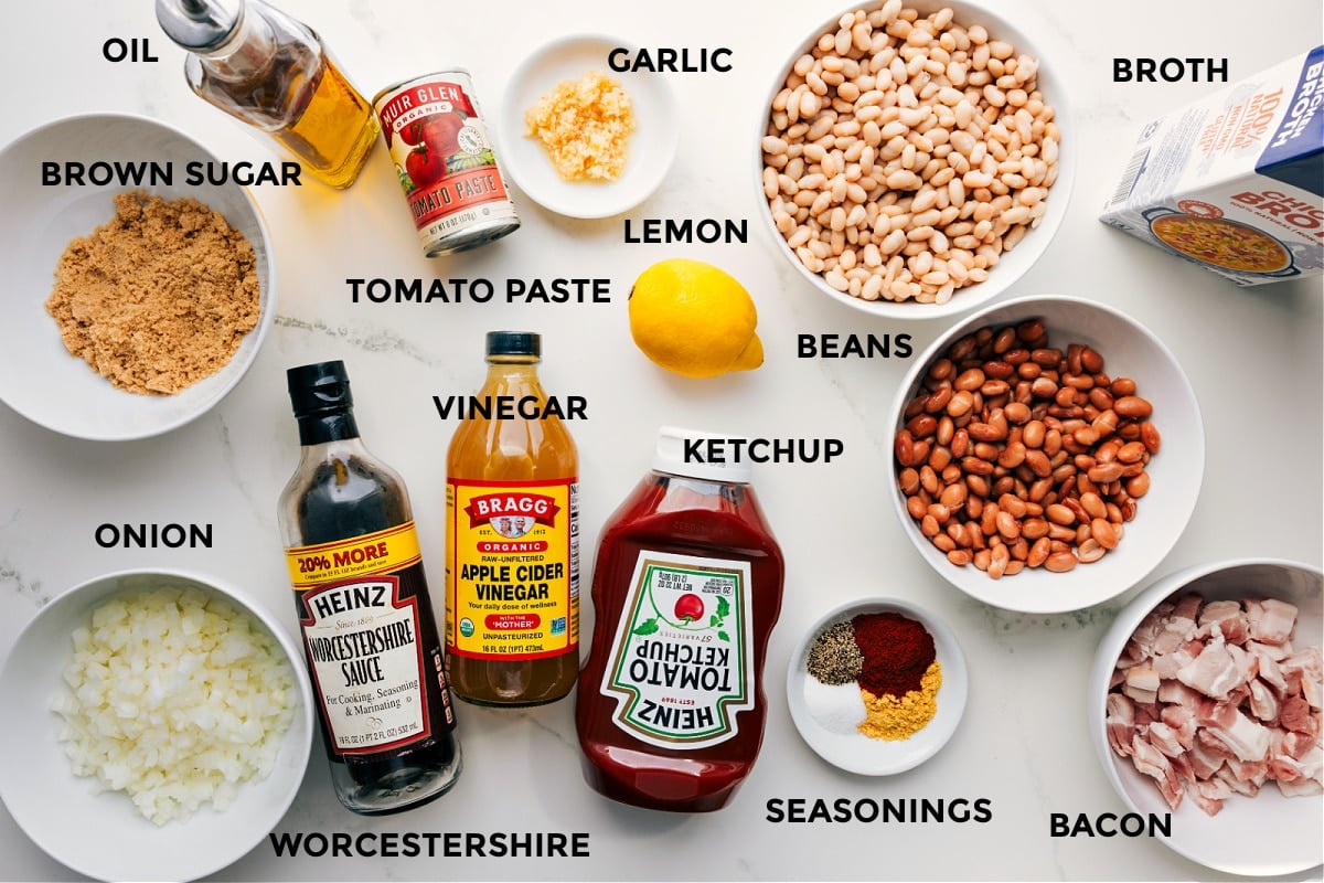 All the ingredients for baked beans prepped and ready to assemble, including oil, brown sugar, onion, tomato paste, vinegar, Worcestershire, garlic, lemon, ketchup, beans, spices, bacon, and broth.