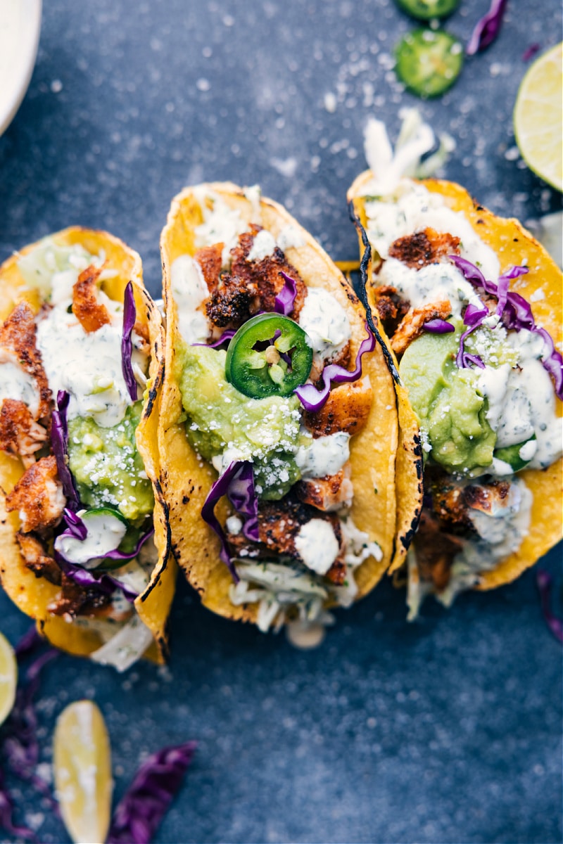 Tilapia Fish Tacos start with charred corn tortillas and are loaded with a saucy cilantro-lime slaw, taco-seasoned Tilapia, and a scoop of guacamole. Recipe via ChelseasMessyApron #dinner #quick #easy #simple #best #fresh #clean #cleaneating #familyfriendly #kidfriendly #tilapia #fish #tacos Overhead image of the tacos ready to be enjoyed