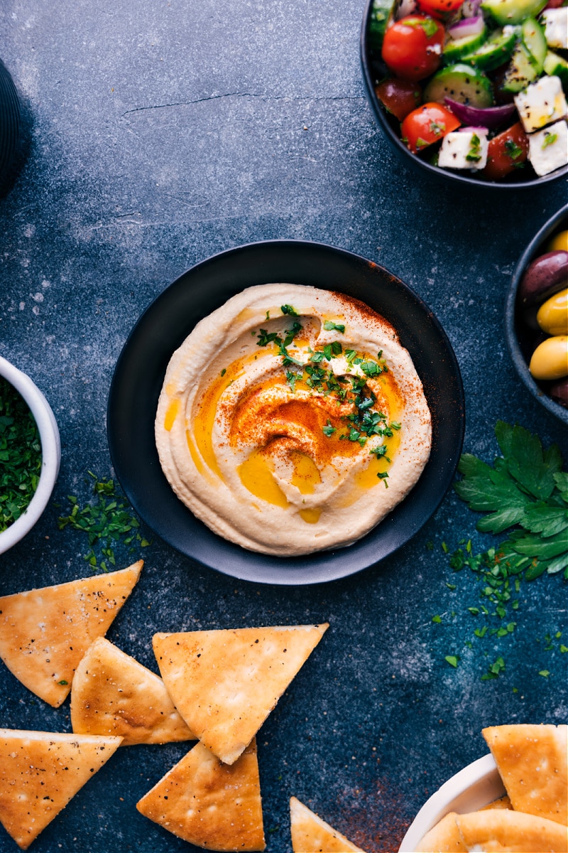 View of a bowl of Hummus surrounded by Israeli Salad and pita chips
