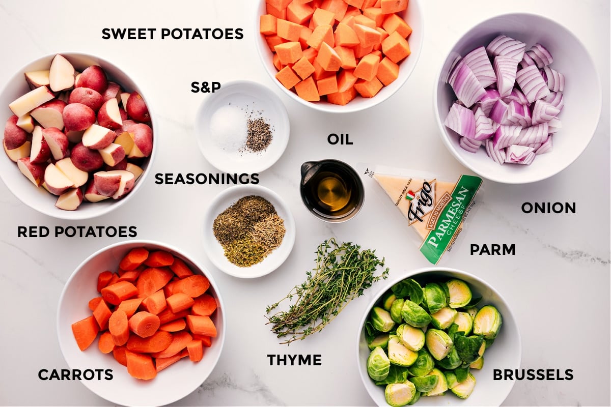 Ingredients In Roasted Vegetables All the ingredients in this recipe prepped out for easy assembly including the potatoes, carrots, sweet potatoes, seasonings, oil, thyme, Brussels, parmesan, and onion,