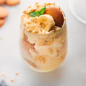 A glass filled with layers of the delicious banana pudding recipe, alternating between pudding, vanilla cookies, fresh bananas, and topped with vanilla cookie crumbs, creating a delightful dessert.