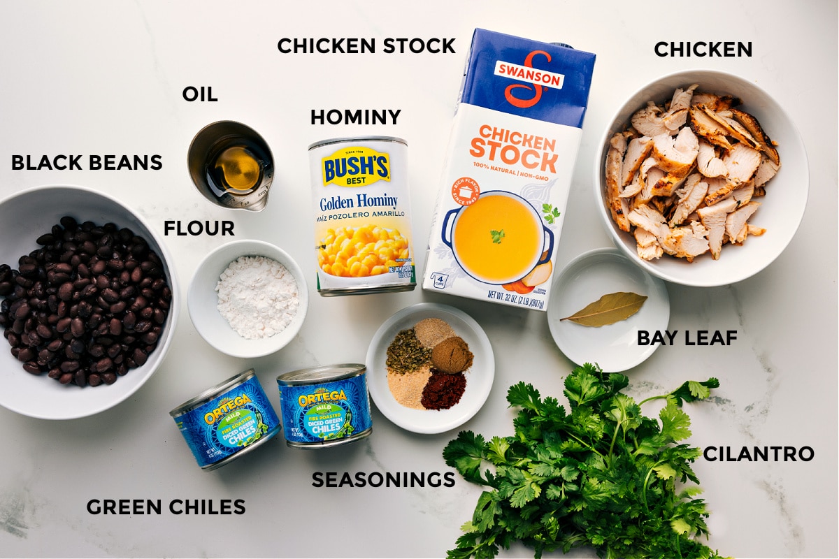 Prepped ingredients for this recipe laid out for easy assembly, including black beans, flour, green chiles, seasonings, oil, hominy, stock, bay leaf, chicken, and cilantro.