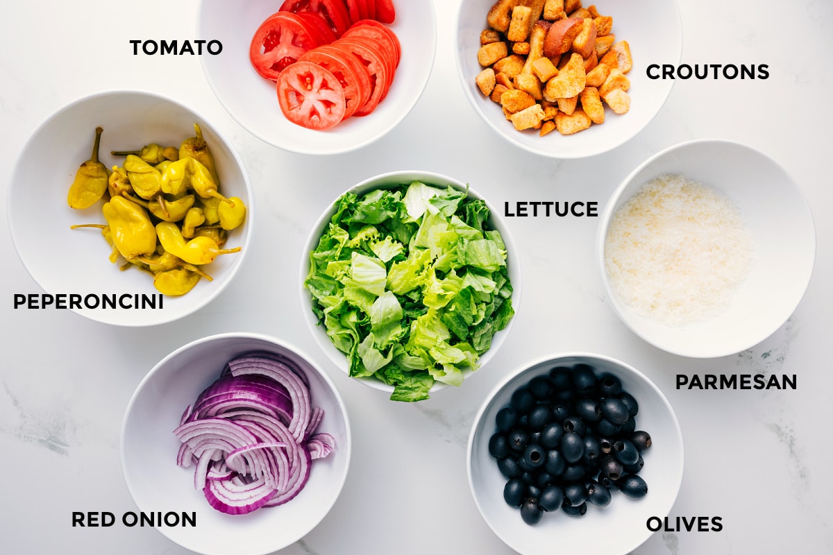 All the ingredients in this recipe prepped out for easy assembly including the peperoncini, red onion, tomato, lettuce, olives, parmesan, and croutons.