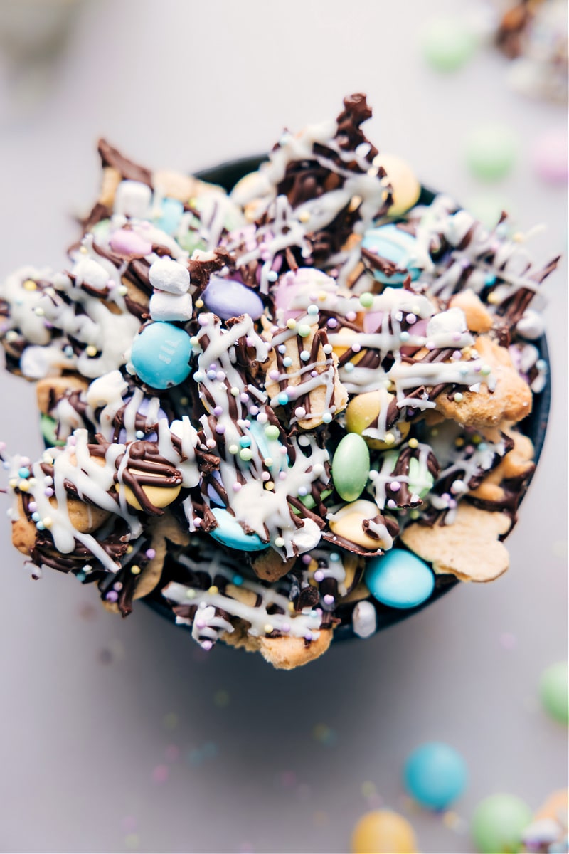 Spring has sprung, and with Easter right around the corner, it's the perfect time to start planning your holiday treats! If you're looking for a fun and festive snack that's both delicious and adorable, then you'll love this Easter Snack Mix. This recipe is inspired by a classic S'mores, with a combination of milk chocolate, Bunny graham crackers, Spring M&Ms, and marshmallows. And the best part? It's topped off with a white chocolate drizzle and Easter sprinkles, creating the perfect finishing touch! Recipe via chelseasmessyapron #dessert #treat #snack #spring #springtreat #easter #eastersnack #easterdessert #eastersnackmix #snackmix Overhead image of the Easter Snack Mix in a bowl