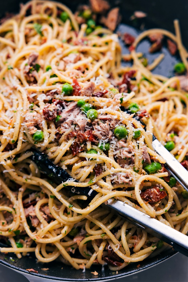 Tongs scooping up a hearty portion of flavorful and healthy tuna salad pasta, garnished with freshly grated cheese, showcasing a blend of vibrant ingredients and fresh appeal.