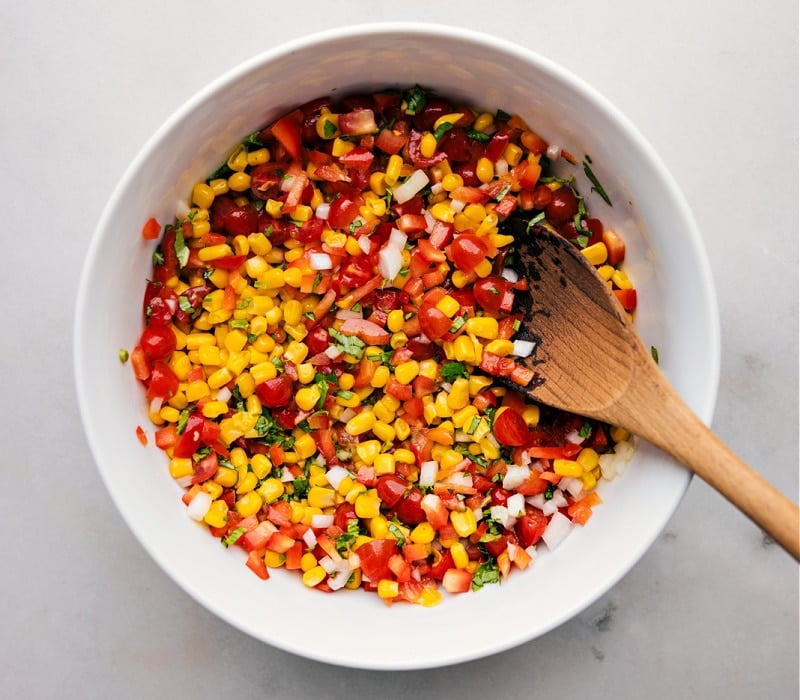 Overhead view of a bowl of Corn and Tomato Salad with a wooden spoon in the bowl.