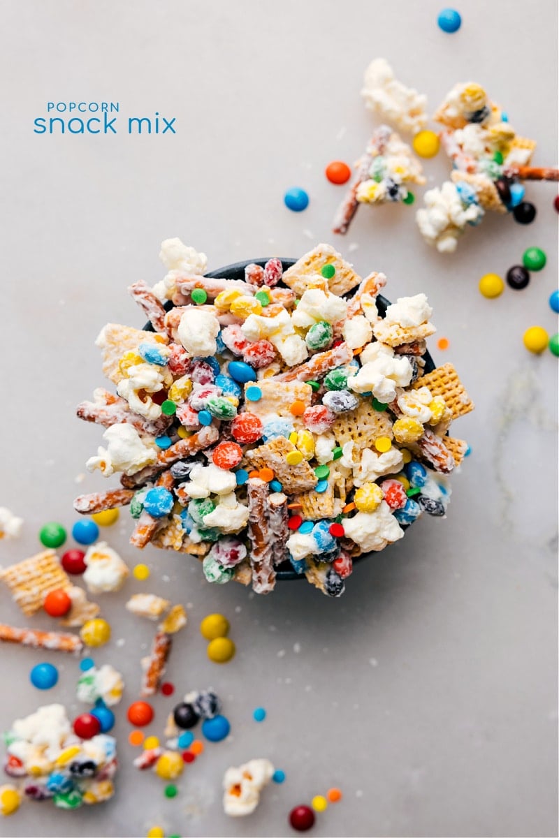 This Popcorn Snack Mix is a tasty mix of sweet and salty elements. It features the crunchy duo of popcorn and pretzels mixed with sweet Chex cereal and mini M&M's, all covered in a layer of creamy white chocolate. Throw in some sprinkles for extra fun and sweetness, and voila - you've got a totally irresistible snack! Best of all, it's simple to whip up with no baking needed! Recipe via chelseasmessyapron #dessert #best #quick #easy #simple #treat #whitechocolate #chexmix #m&ms #popcorn #snackmix Overhead image of the Popcorn Snack Mix ready to be enjoyed