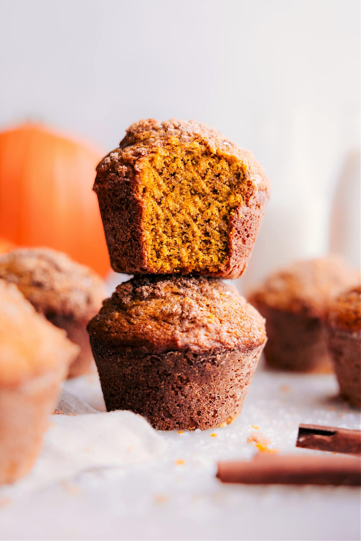 A stack of pumpkin muffins with a warm, golden tone, one at the front displaying a bite taken, revealing the moist interior.