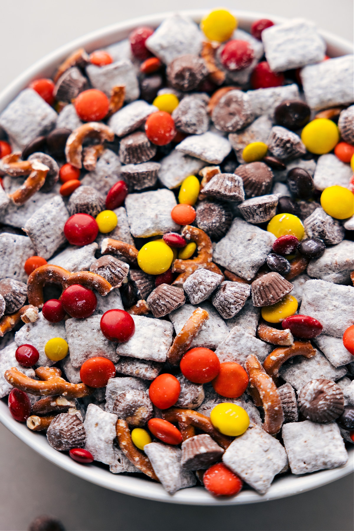 A bowl filled with Fall Muddy Buddies, showcasing the enticing mixture of chocolate-coated, powdered sugar-dusted cereal, punctuated with vibrant, seasonal mix-ins for a perfect autumn treat.