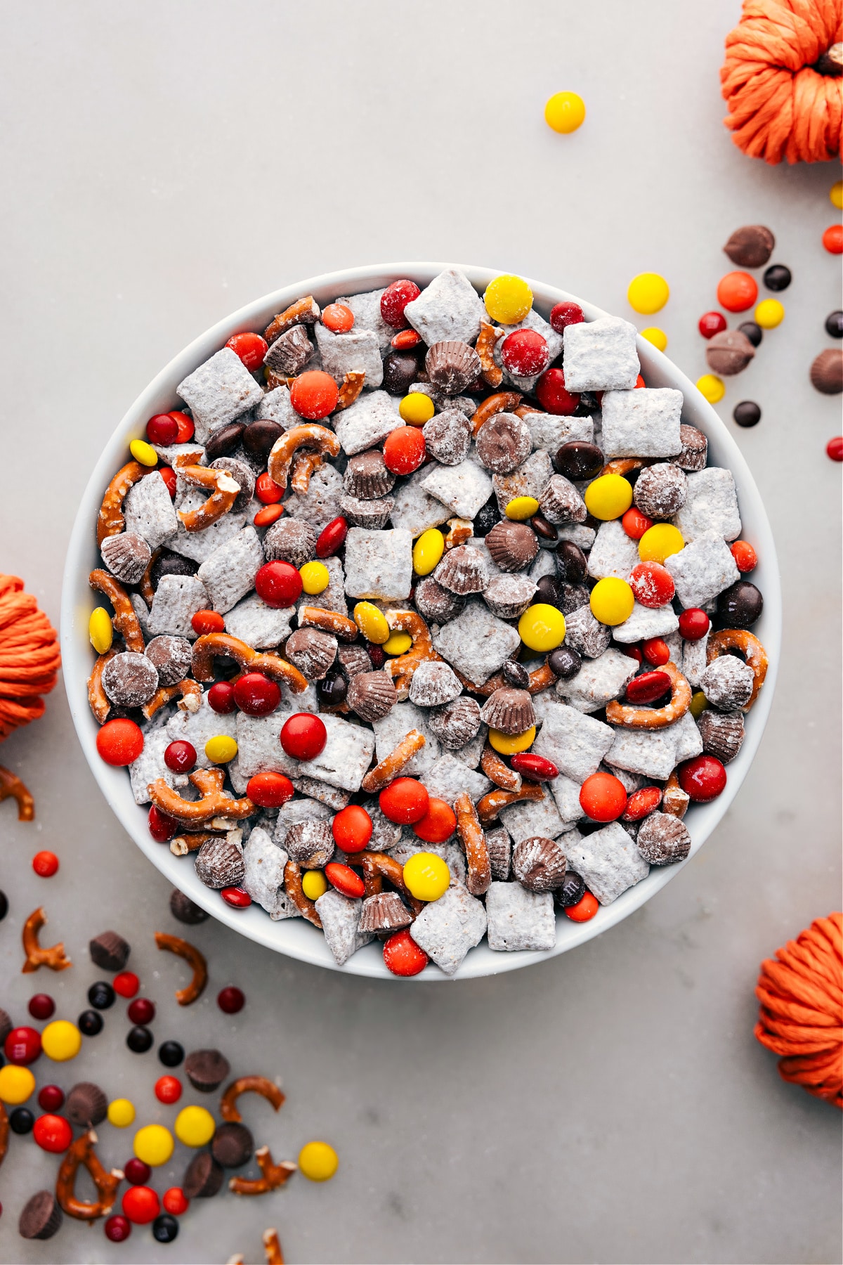 A bowl of Fall Muddy Buddies, showcasing the delightful mix of cereals coated in chocolate and powdered sugar, punctuated by autumn-themed candies and snacks for a festive treat.