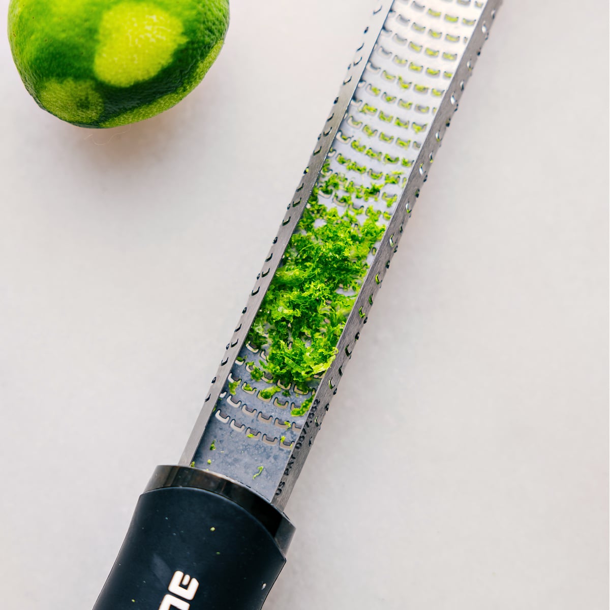 A zester tool covered in fine lime peel shavings, highlighting the fresh, green zest just removed from the fruit, ready for use in this recipe.