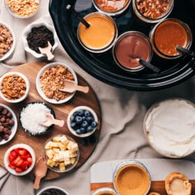 Various dessert toppings and fillings including glass jars filled with caramel, chocolate, and other sauces, accompanied by bowls of crushed nuts, blueberries, coconut shreds, chocolate chips, and other delectable ingredients for a dessert pie bar.