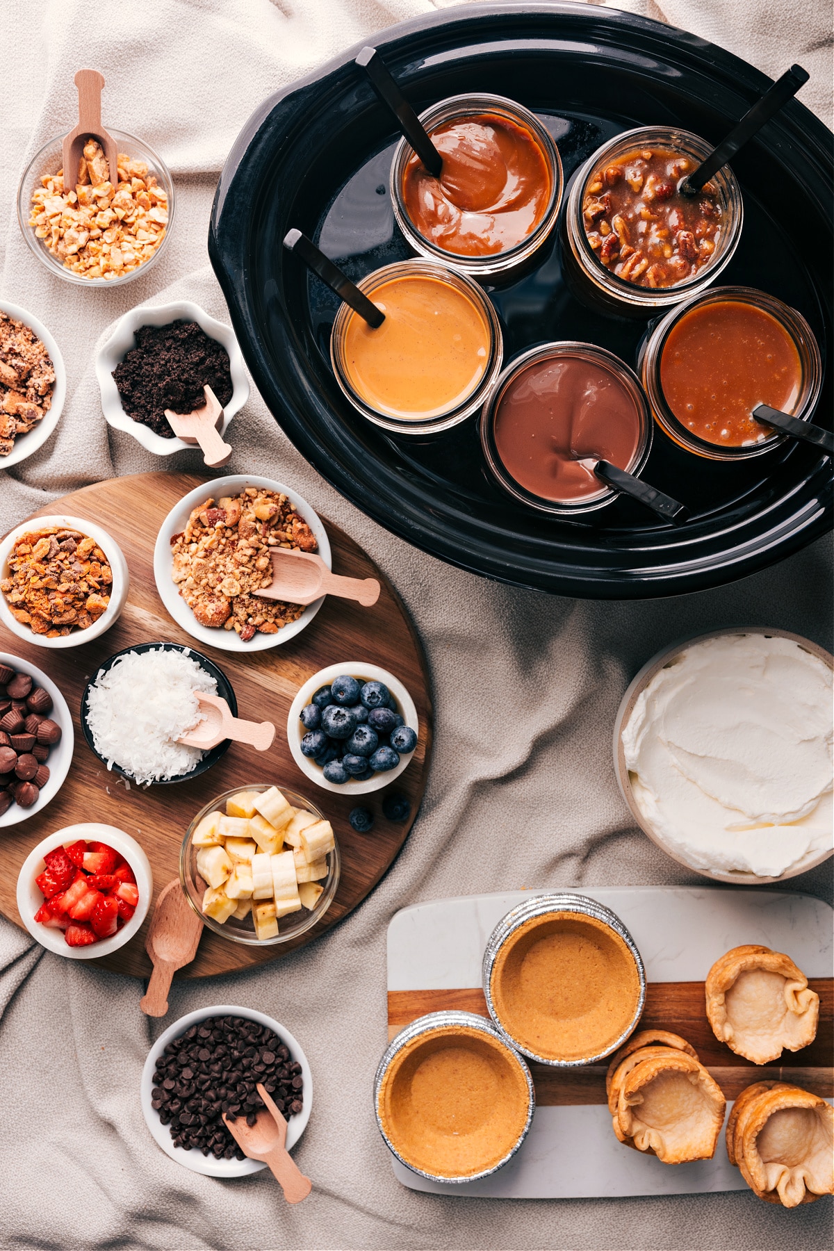 Various dessert toppings and fillings including glass jars filled with caramel, chocolate, and other sauces, accompanied by bowls of crushed nuts, blueberries, coconut shreds, chocolate chips, and other delectable ingredients for a Dessert Pie Bar.