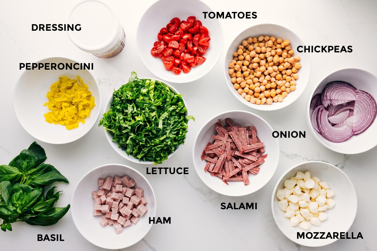 Italian chopped salad ingredients prepped for easy assembly, including dressing, pepperoncini, basil, ham, lettuce, tomatoes, salami, chickpeas, mozzarella, and onion.