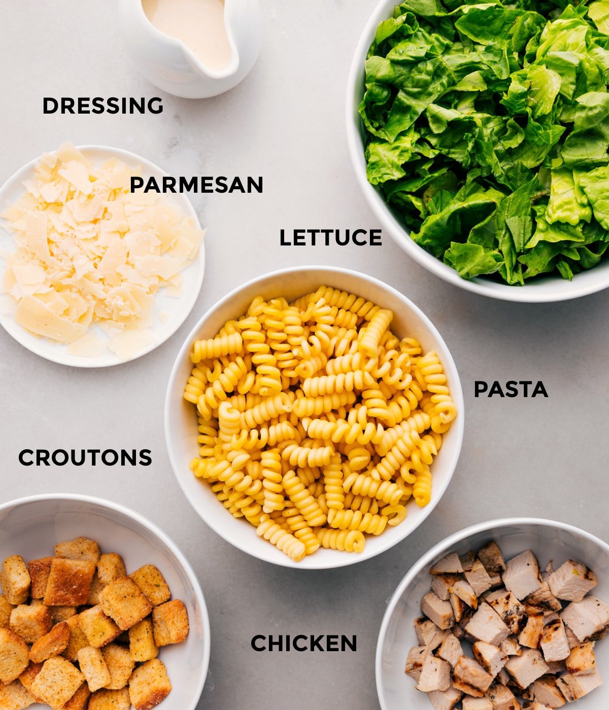All the ingredients for this recipe prepped and ready for easy assembly, including the dressing, parmesan, lettuce, pasta, chicken, and croutons.