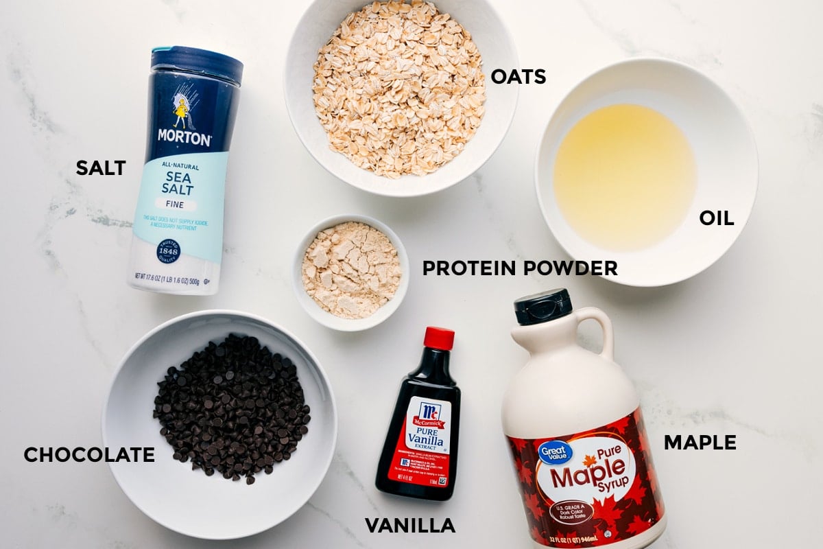 All the ingredients for this recipe, prepped and ready for easy assembly: oats, oil, salt, protein powder, vanilla, maple syrup, and chocolate.