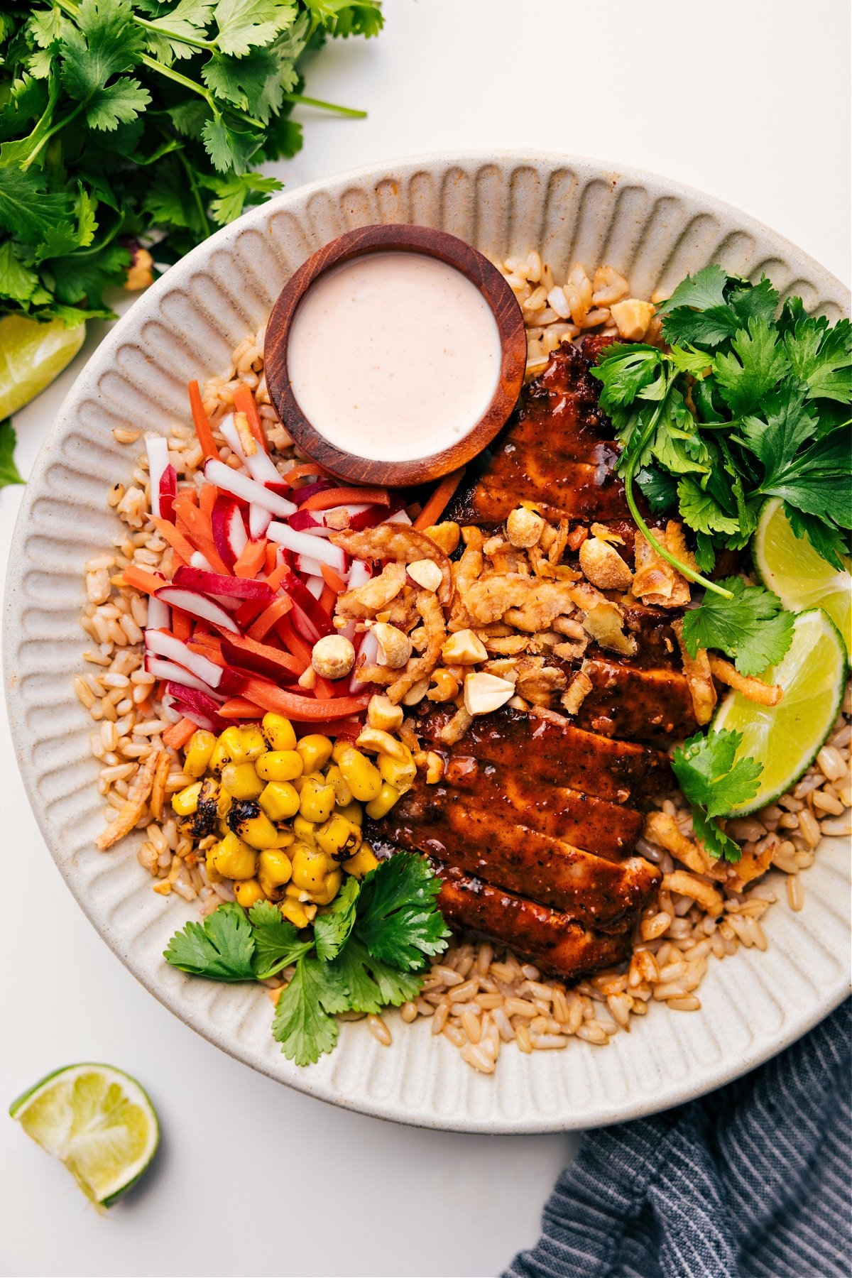 Asian Chicken Bowl served on brown rice, topped with corn, radish and carrot salad, limes, cilantro, and peanuts, with sauce on the side.