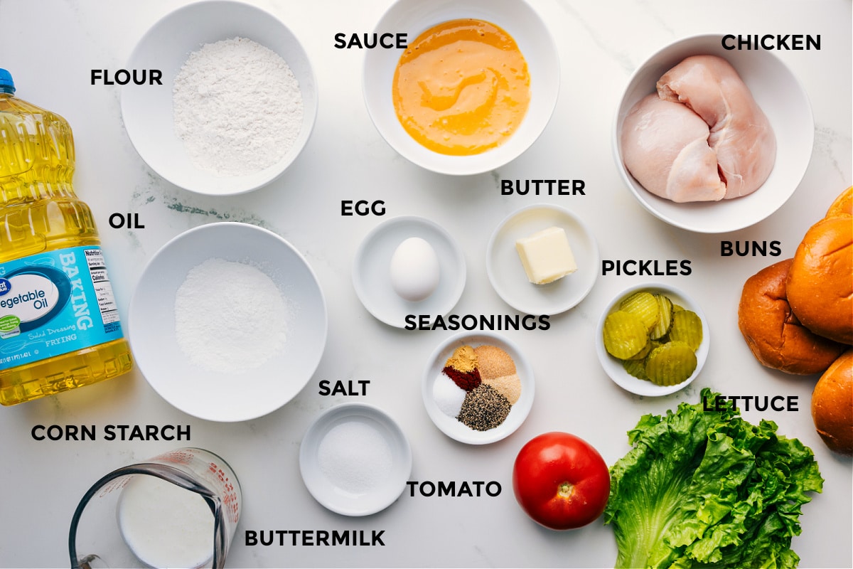 All the ingredients prepped and ready for crispy chicken sandwich assembly, including oil, cornstarch, flour, sauce, egg, seasonings, salt, tomato, butter, pickles, lettuce, chicken, and buns.