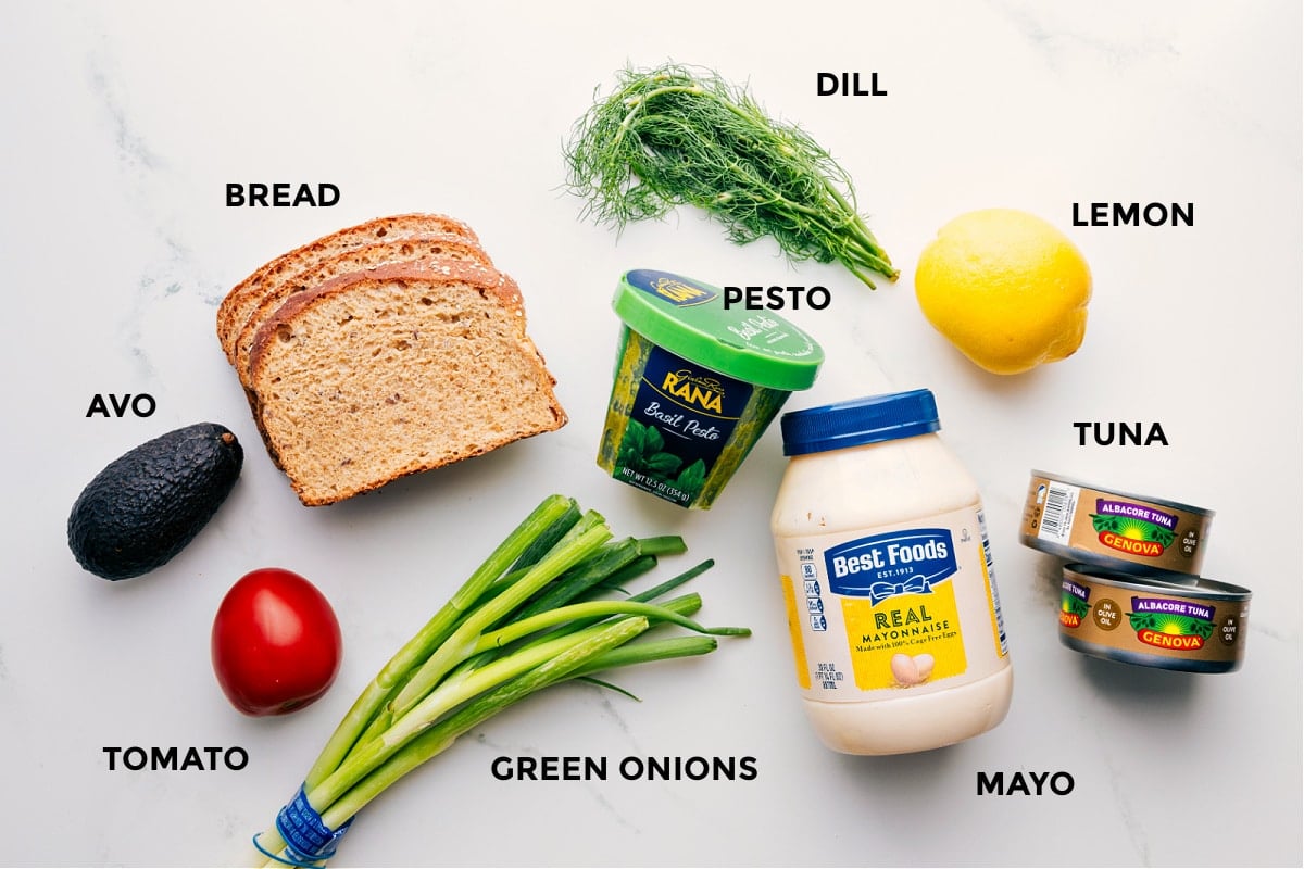 All the ingredients prepped for easy assembly, including avocado, tomato, bread, green onions, pesto, dill, lemon, tuna, and mayo.