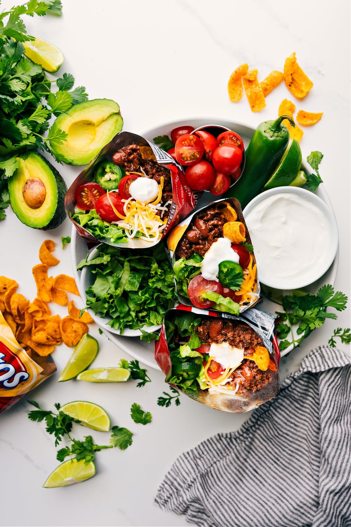 Summer Must Have: Walking Tacos Walking Taco Recipe served on a plate with three chip bags filled with saucy ground beef, sour cream, tomatoes, lettuce, and jalapeños.