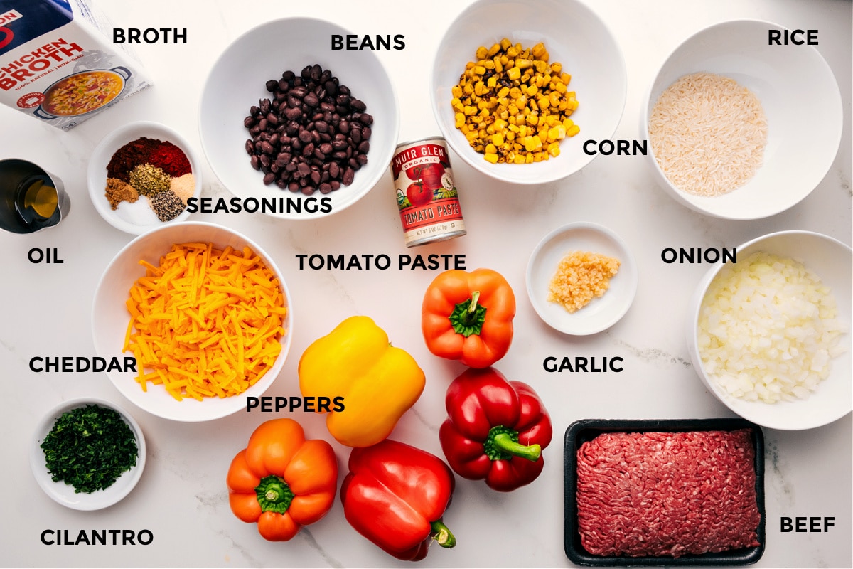 Stuffed pepper ingredients prepped for easy assembly, including broth, oil, spices, cheddar cheese, cilantro, peppers, beans, tomato paste, garlic, ground beef, rice, corn, and onions.