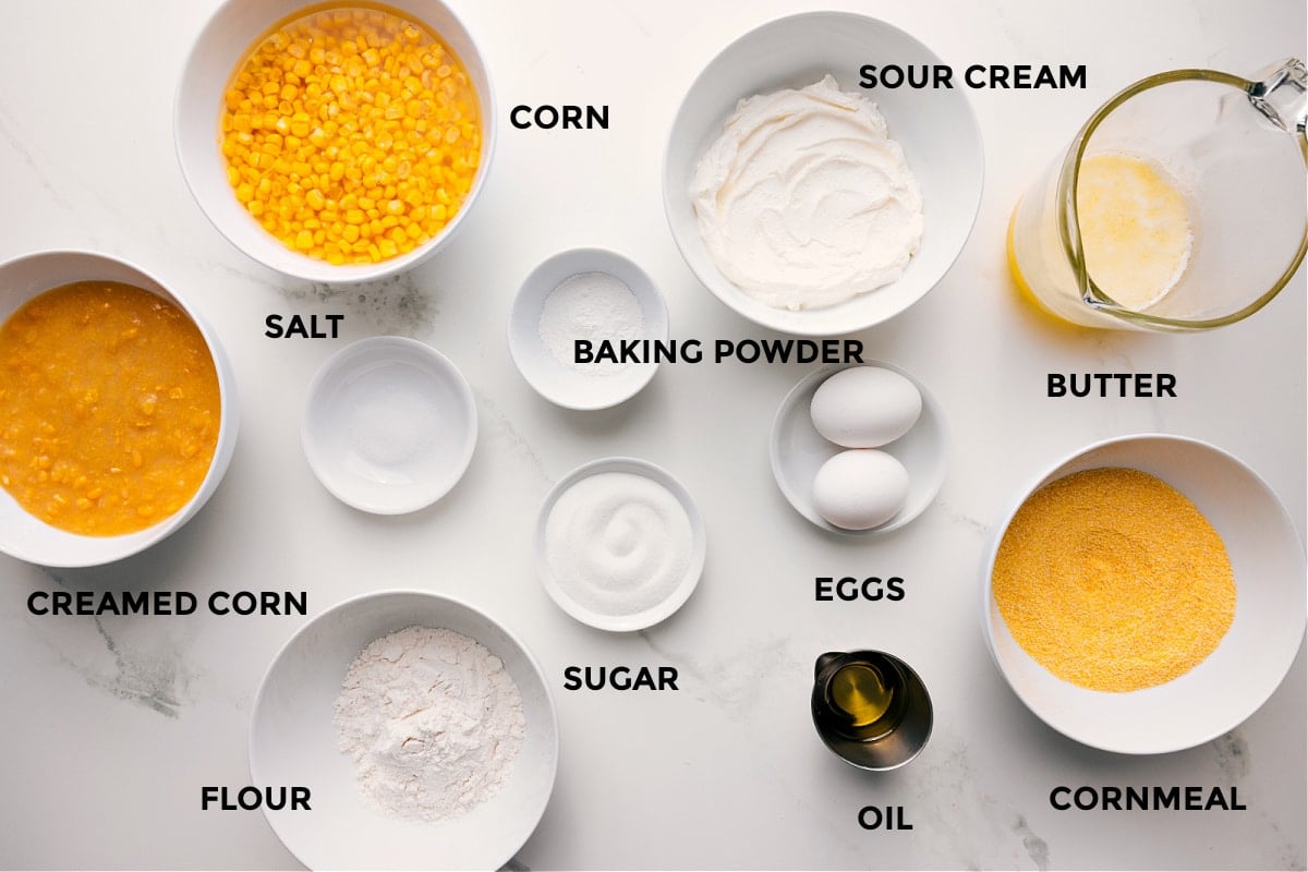 All the ingredients in this recipe prepped out for easy assembly including the creamed corn, corn, salt, flour, baking powder, sugar, sour cream, eggs, oil, cornmeal, and butter.