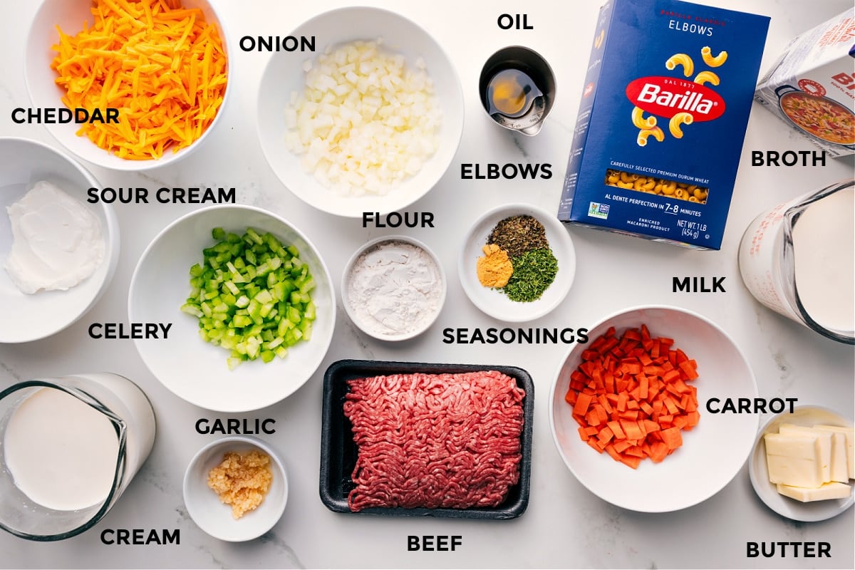 All the ingredients for this recipe prepped and ready for easy assembly, including sour cream, cream, cheddar, onion, celery, garlic, flour, ground beef, seasonings, oil, carrot, milk, butter, pasta, and broth.