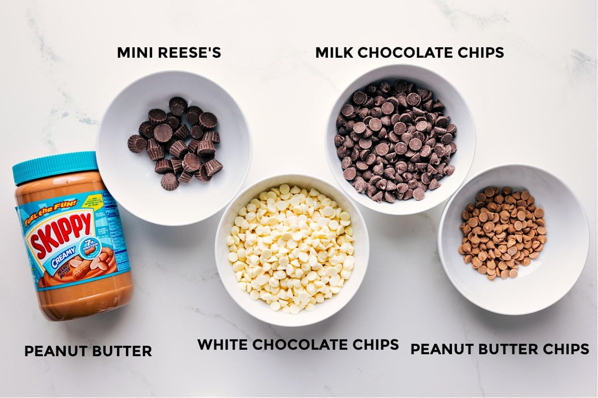 Ingredients In Peanut Butter Bark Ingredients prepped in small bowls for easy assembly including peanut butter, mini Reese’s cups, white chocolate chips, milk chocolate chips, and peanut butter chips.