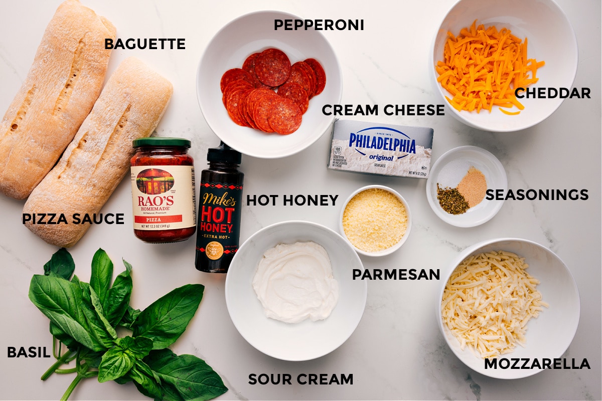 Ingredients In Pizza Dip Recipe All the ingredients in this recipe prepped out for easy assembly including the baguette, basil, pizza sauce, hot honey, sour cream, pepperoni, cream cheese, parmesan, mozzarella, seasonings, and cheddar.