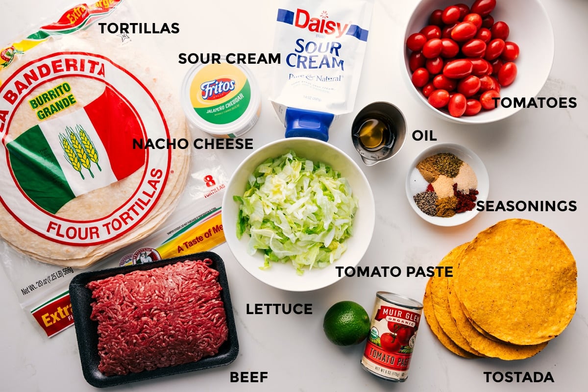 All the ingredients in this recipe prepped out for easy assembly including the tortillas, beef, lettuce, tomato paste, lime, seasonings, tomatoes, oil, sour cream, and nacho cheese.