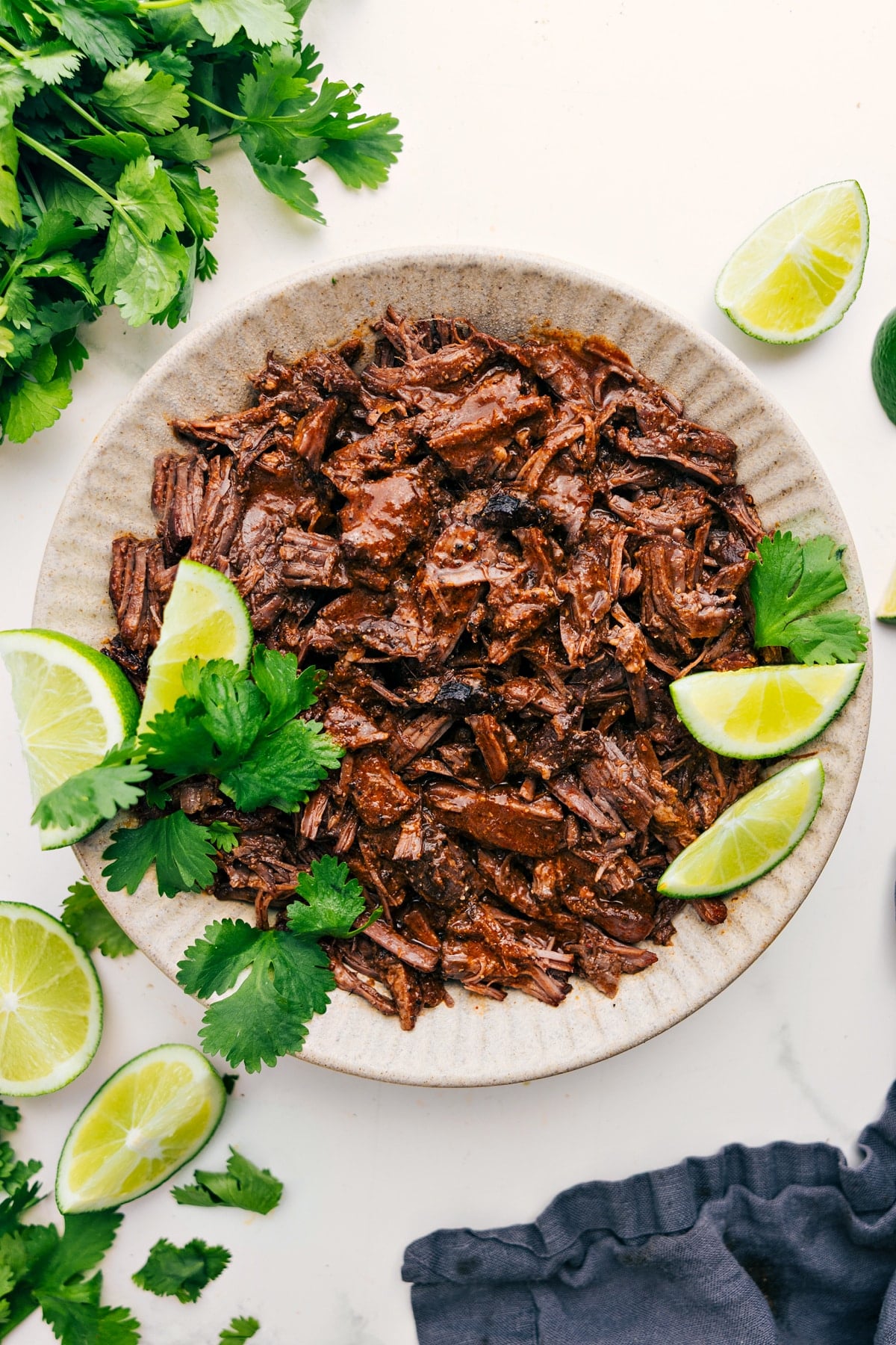Shredded Barbacoa Beef Recipe Barbacoa Beef Recipe in a bowl fresh from the slow cooker with lime wedges and cilantro on the side.
