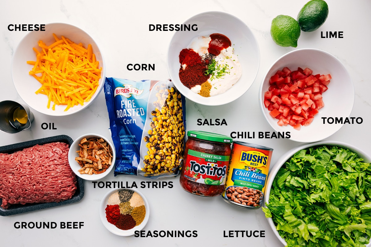 Ingredients In This Taco Salad Recipe All the ingredients in this recipe prepped out for easy assembly including the ground beef, oil, cheese, tortilla strips, seasonings, salsa, chili beans, lettuce, tomato, lime, and dressing.