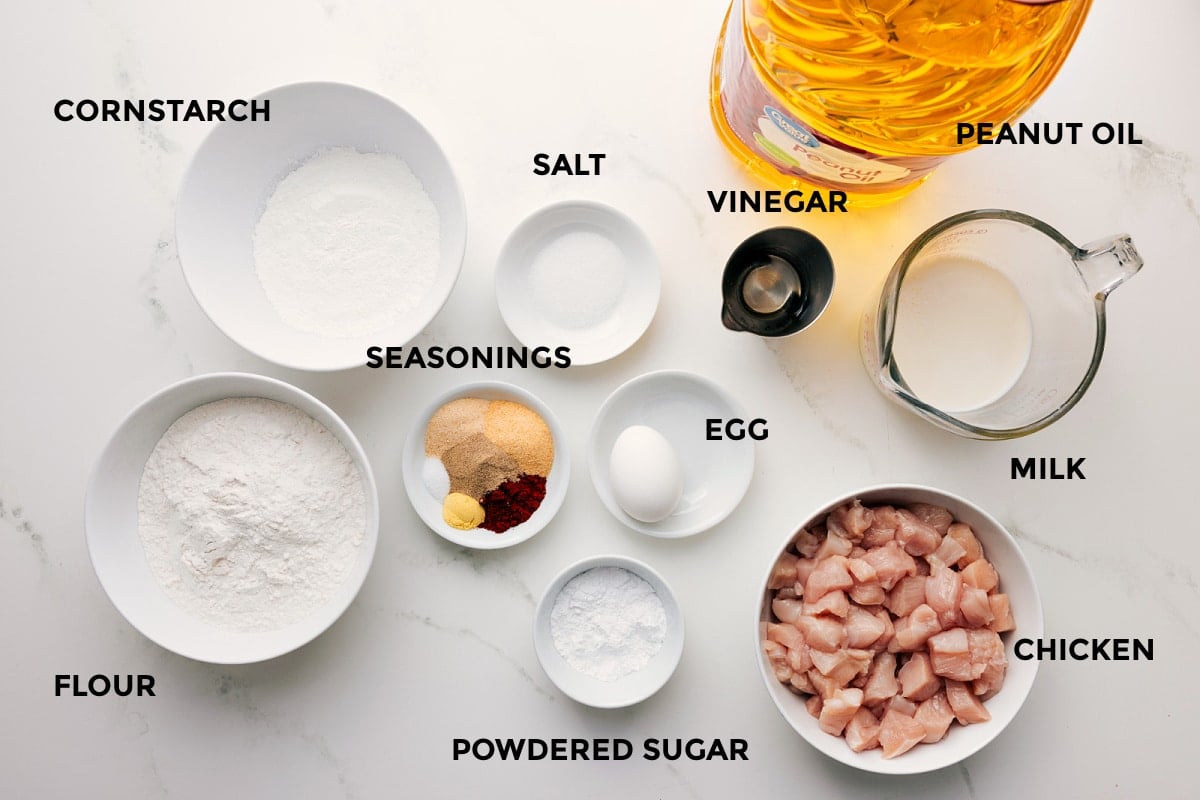 All the ingredients in this recipe prepped out for easy assembly including the cornstarch, flour, seasonings, salt, powdered sugar, egg, vinegar, peanut oil, milk, and chicken.